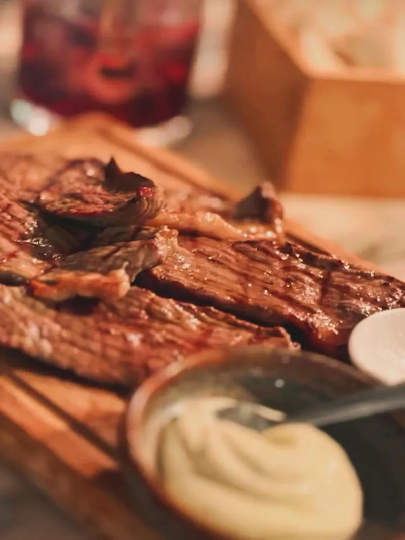 Fatias de carne grelhada numa tábua de madeira, com um molho branco ao lado.